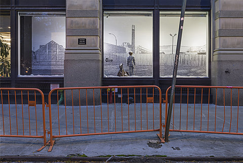 A window viewed from the street displaying a small child in pajamas facing a wall in a snowy scene alongside a small dog peeking out of a bag. Three orange barrier railings are in the foreground.