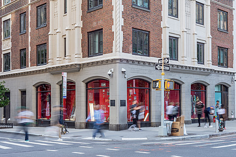 The corner of a building seen from street-level. People walk in front of ground floor windows filled with various items against red backgrounds.