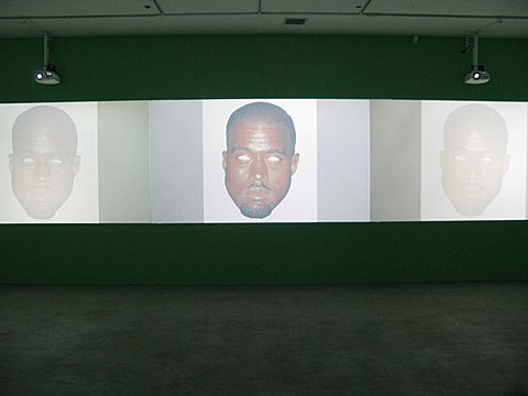 There is a green wall with three projected images of Kanye West’s face on a white background side by side. Beside the white background of the images are thick tinted boarders on the right and left side of each image.  Kanye West’s eyes in each image are missing and instead all we see is the white background where his eyes would be. 