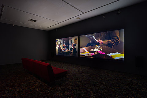 Red couches sit in front of two large projections showing people sitting around a small table.