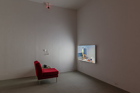 A red seat sits in front of a TV monitor which displays a person painting. Headphones rest on the seat. A white piece of paper with text is taped to the wall.
