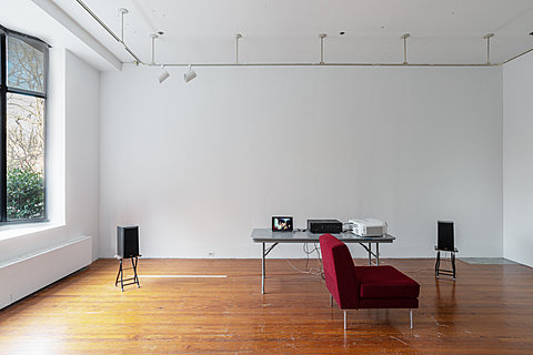 A red couch sits perpendicular to a grey table, which holds a laptop, stereo reciever, and projector. Two speakers flank the table.