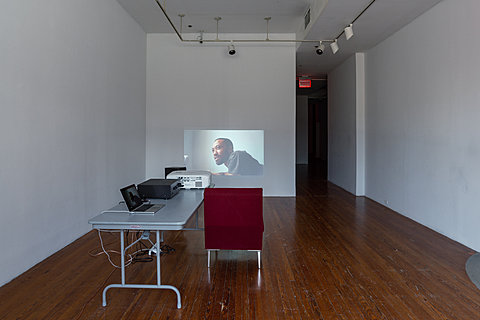 A red couch faces a wall onto which is projected a close-up shot of a person mid-conversation.