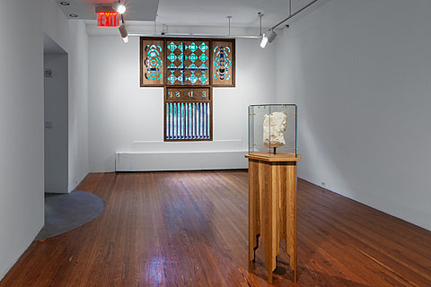 A carved stone fragment of an animal head is displayed on a wooden pedestal, sealed by glass sheets with metal fittings. In the background is a window covered by a wood cutout.