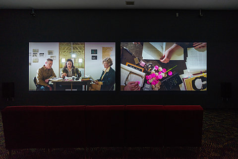 Red couches sit in front of two large projections showing people sitting around a small table.