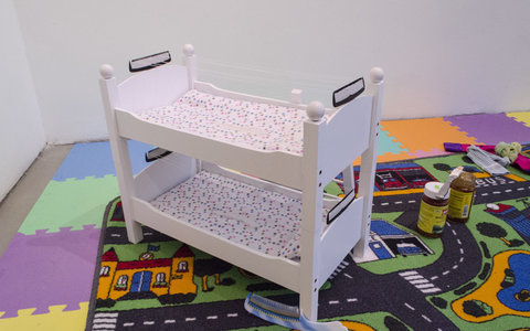 Close-up image of the installation featuring a multi-colored play mat made of foam. On top of the mat is a small rug with cartoon roads, and town on it. Sitting atop the rug, is a doll-house sized, bunk bed toy made of white wood. 