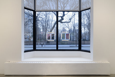 Installation view of the exhibition featuring the window in the gallery with four panels of glass and two artworks hung in the two center panels. In the left window pane, two abstract prints are taped to the glass and on the right side, two other prints are taped to the glass.