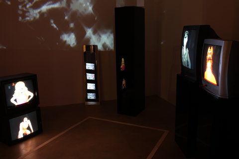 Installation view of the exhibition featuring a dark room with a projection on one wall. The projection is abstract, dark and blurry. There are several television monitors stacked on top of one another and placed throughout the room with various figures on the screens.  