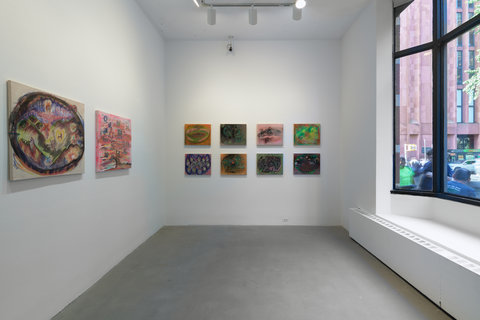 Installation image of several works in the exhibition. On the left, two paintings hang against a white wall. On the back wall two rows of four works of art hang on the back wall, evenly spaced apart. On the right, a large window lets lots of light into the space. 