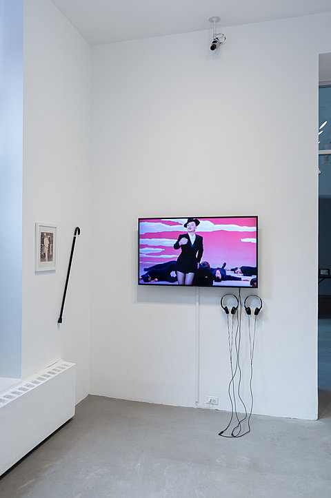 A television monitor with two headphones hanging on the wall is displayed next to a black cane and framed image.