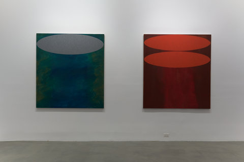 Installation view of two works hanging on a white wall in the exhibition. Each work is slightly illuminated by a white spotlight. The work on the left, is painted with mainly blue hues, the subject is abstract and features a light grey oval painted at the top of the painting. The painting on the right features mostly red hues, with two red ovals painted on top of one another at the top of the canvas. The white wall is contrasted by the cement floor of the gallery in the foreground. 