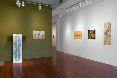 Installation view of three works in the exhibition. The wall is painted dark olive green. The work features a drawing on a scroll that is placed on a shelf halfway up the wall. The scroll falls down to the floor, revealing a line design of two thick intertwining green lines with small yellow lines drawn inside it. The work on the right features a thin, undefined, green tree on a hill of dark brown and light orange/brown patches of dirt. White lines protrude from the bottom of the tree throughout the hill, resembling roots.  On the opposite wall, which is painted white, three drawings hang on the wall, lit up individually by spotlights. 