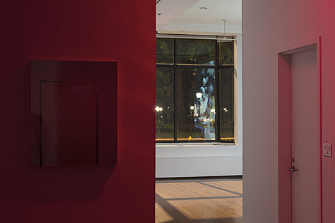 Shot from a room filled with red light, a window can be seen in the distance with the reflection of a wall projection visible in it.
