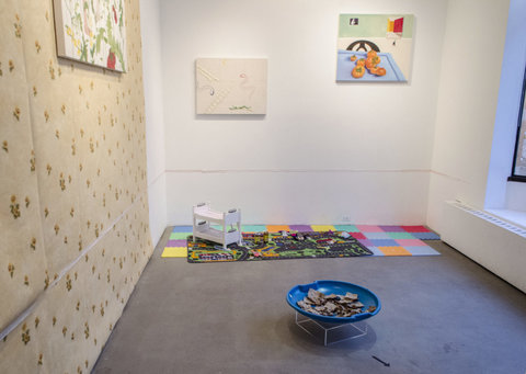 Installation view of the exhibition featuring several artworks in the exhibition. A blue bowl sits in the center of the gallery, on the cement floor with ripped up pieces of brown paper inside. The left wall is covered in floral wall paper. Several paintings are hanging. in front of the middle wall, a play mat is set up. It is made of mutli-colored patches of foam and includes a rug that has roads and foliage on it. There is a small white, toy-sized bunk bed on the carpet.  