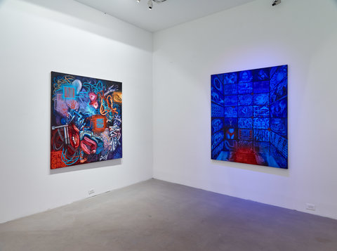 Installation image of two walls. On the left wall is a large square painting featuring many abstract shapes and colors. On the right is a large blue and red square painting that is made up of smaller squares in different shades of blue featuring different images in each square. The canvas looks as though it is glowing. 