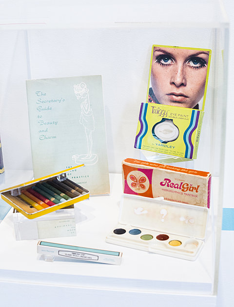 A display case holding 1960's makeup. In the background is a book titled "A Secretary's Guide to Beauty and Charm."