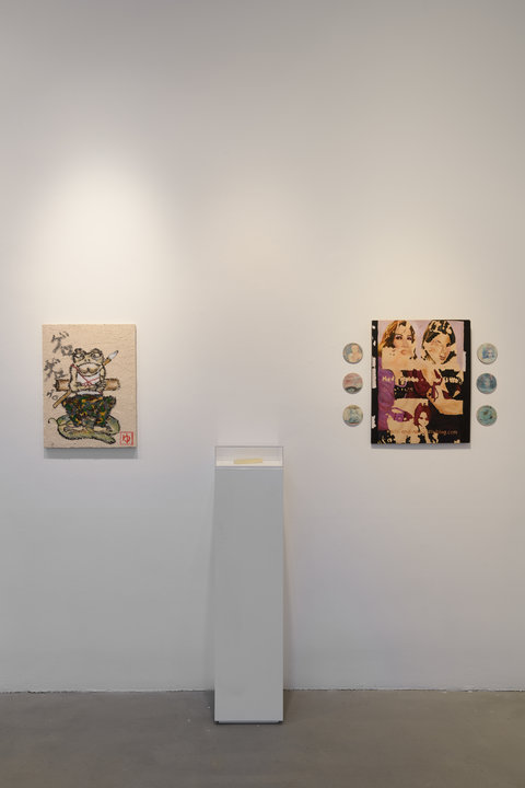 Installation view of three objects in the exhibition against a white wall. The first object is a painting with an anthropomorphic frog figure crossing its arms and wearing clothes as it balances a top a lily pad. Next to this frog, a white pedestal sits against the wall with a small glass box on top. The contents of the pedestal are illegible. On the right side of the pedestal, a painting hangs with three circular discs on each side.