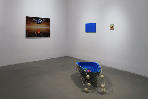 In the corner of this gallery room there is a kiddie pool-like sculpture sitting on the floor. The sculpture has a wooden frame around, and rubber duckies floating around the water inside of the pool. On the wall to the right of the sculpture there is a small painting of a cat, and to the left of the cat is a larger blue painting. On the left wall is a large woven textile piece that has been stretched over a frame to hang on the wall. This piece features an upside volcano-like form. 