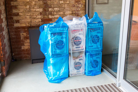 Installation view of an object from the exhibition. The structure consists of six boxes stacked on one another in three by two stack. The first two are wrapped in transparent blue bags, the middle boxes are in transparent white bags, and the last two are in transparent blue bags. 