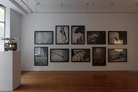 Ten black and white photographs fill up a white wall in a room with a wood floor. On a pedestal to the left of the photographs is a small metal projector.    