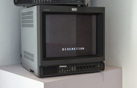 Image of a television monitor in the installation featuring a black screen with the word "DISCRETION" on the bottom. The monitor sits atop a white pedestal. 