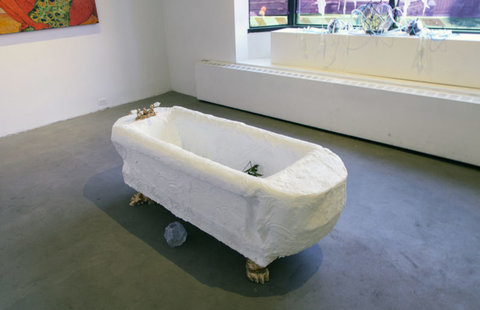Installation view of the exhibition featuring a bathtub structure in the center of the gallery floor. The tub is made of what seems to be paper mache. 