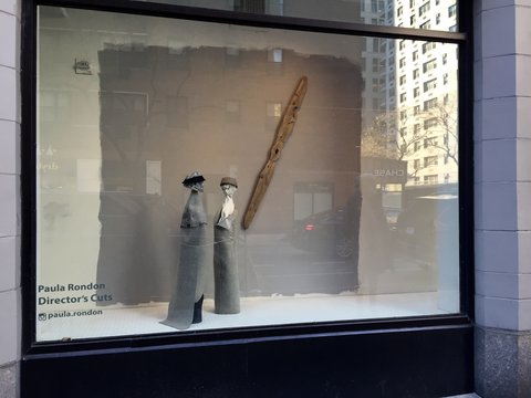 Image of the exhibition from street view looking into the window from the outside. The window installation features two sculptures sitting atop grey carpet like pedestals. The sculptures resemble heads with headbands on, perhaps even skull like. The grey carpet is also attached to the wall in the background. On the back wall, a long diagonal piece of wood is mounted to the carpet. On the window, the words, "Paula Rondon, Director's Cuts" are printed onto it. Below those words, the instragram logo appears, with the handle "paula.rondon". 