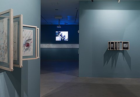 A framed series of graphite drawings are hung on a blue gallery wall. In the background can be seen a room with a projection on the wall.