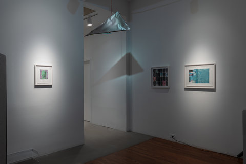 Installation view of three framed artworks in the exhibition. On the left wall, an image in a white frame is spotlighted by a light from above. On the left wall, the left most image is not spotlit, but the right image, that is mostly blue, is lit with a spotlight. In the middle, hanging from the ceiling is a metallic triangular mobile that reflects a strong shadow behind it. 