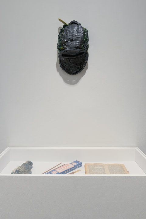 In this photo we see a display case and a ceramic hat-like form hanging on a wall. In the display case there are three objects. On the left there is an oblong rounded ceramic form. In the middle there is what appears to be a box of fire sparklers. On the right there is a book sitting open. The steal-colored ceramic hat hangs above the display case.