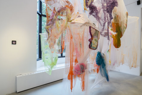  In this gallery room there is a large installation of mosquito nets, tule, and foam cutouts hanging from the ceiling. The mosquito nets are torn in some places and tied together in others. The bottoms of these nets are painted with colors such as purple, blue, green, and orange. On the foam cutouts are colorful portrait drawings. On the window wall behind the installation there is a very small frame. This thick black frame is to the right of the window and holds a photograph of a woman with a purple tint. 
