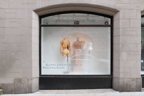 Image of three mannequins in the exhibition. The mannequin on the left is faced away from the audience and wears a bag of some sort draped over its shoulder. The mannequin on the left is legless, and headless. The mannequin in the middle wears a pink, tulle corset top with flowing sleeves, and a slim light pink skirt. On the right, the mannequin is only a torso and includes a white corset top. 