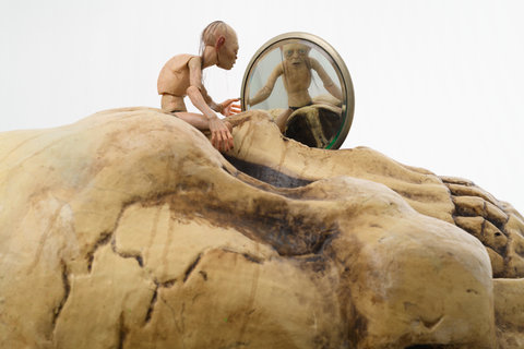 Close-up image of a sculpture work. The work features a small Gollum figurine from the Tolkien series. The figure is sitting atop an oversized skull which is facing up towards the ceiling. The figure looks into a small pocket mirror and it's reflection stares back at the viewer to reveal open eyes and strings of hair.  