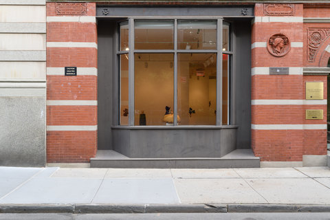 Image from outside the building featuring a large glass window with two main front facing panes. One can see outlines of the objects within the exhibition. The building's red and white striped brick exterior frames either side of the window. 