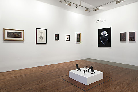 Eight framed artworks hang on a gallery's walls, each in a different frame. A short pedestal on the ground has several sculptures of birds with human faces.