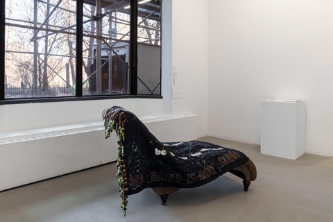Installation view of the exhibition featuring a sculpture in the middle of the floor. It appears to be a dark brown lounger chair covered by a large black piece of textile with beads and the text “MY AUNT IS AUNT SARAH”.