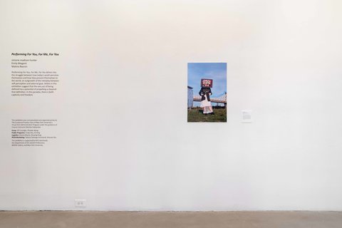 Installation view of one wall in the exhibition featuring the exhibition title “Performing For You, For Me, For You” and exhibition description, and a photography print of what appears to be an outdoor scene of a human figure wearing a rectangular box as a hat, standing on grass. Blue sky covers the background, with a bridge in the background.