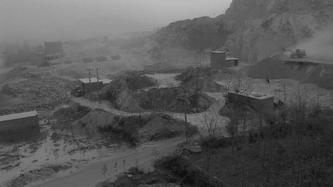 Black and white photograph of an old materials digging site. There is a large pit in the center with mountains surrounding it. There is one tractor on the upper right corner of the image. A fog is present throughout. 