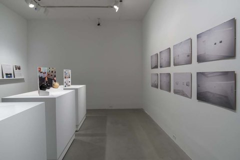 Installation view of the exhibition. On the left, three white pedestals sit with pamphlets atop. On the right, several black and white photographs are mounted to a white wall. 