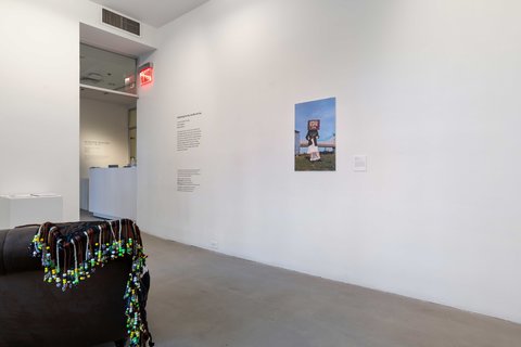 Installation view of the exhibition featuring two walls, a sculpture in the foreground that appears to be a dark brown lounger chair covered by a large black piece of textile with beads and text, and a photographic print of what appears to be an outdoor scene of a human figure wearing a rectangular box as a hat, standing on grass, Blue sky covers the background, with a bridge in the background. 