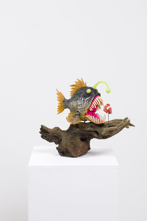 Up-close image of a sculpture featuring the front of an angler fish atop a large, thick branch, on a pedestal. In front of the angler, is a small clown fish.