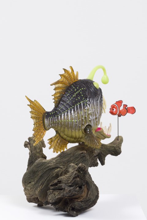 Up-close image of a sculpture featuring the back of an angler fish atop a large, thick branch, on a pedestal. In front of the angler, is a small clown fish.
