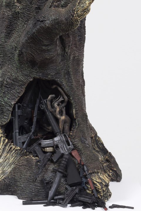 Up-close image of a tree trunk figurine. Various guns and rifles spill out of a hole in the tree. 