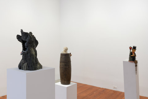 Installation view of three figurines on three white pedestals. On the right, is a figurine of a dark, rotten tree trunk. In the middle is a figurine of a barrel, and on the left, is a figurine of a seated figure holding a change of pennies. 
