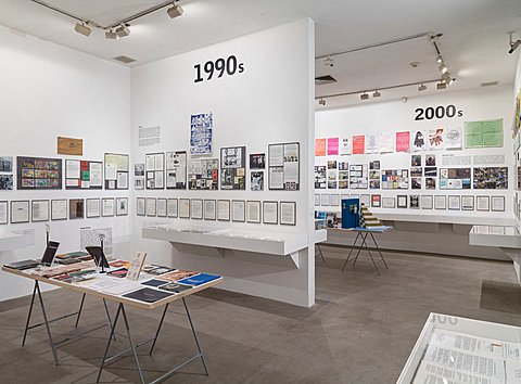 We see half a view of the room showcasing printing media from the 1990s and half of the room showcasing printing media from the 2000s. The 1990s room is seen first and showcases a table in its center that has printed papers placed on its surface. The walls are lined with framed texts and images. The 2000s room behind also has a table in its center and walls lined with framed printed media.