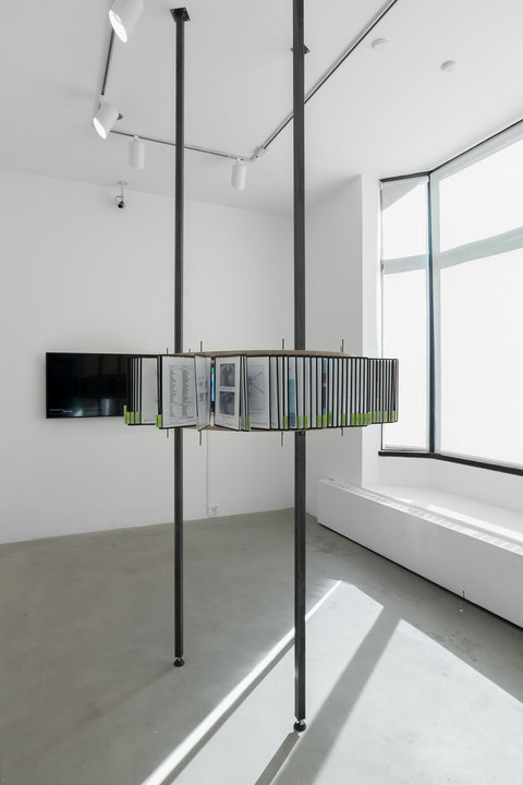 Installation image of the exhibition featuring two tall standing poles in the center of the room. Surrounding the poles, are a series of interactive articles and papers part of a flippable archive. On the right side, large windows pour white light into the room.
