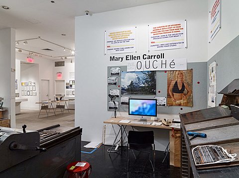 An image and text spread on Mary Ellen Caroll is on a wall above a desk with a desktop computer. On the parallel and right perpendicular wall to the desk there is are old deconstructed printing presses.  
