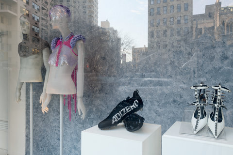 Image of two mannequins in the exhibition and two pairs of shoes. On the left, a mannequin wears a grey hood, and nothing else. The mannequin is missing the lower half of the body. Next to this mannequin, another mannequin wears a pink and purple crocheted item around their head and shoulders. In the foreground, through the window, a pair of black sneakers sits on a pedestal next to a pair of white heels with silver spikes along the straps. 