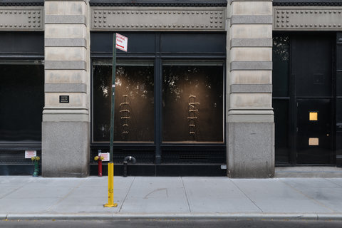 Street view of a light colored building with a large dark window. Inside the window on the left and right sides are 6 short horizontal warm toned neon lights.