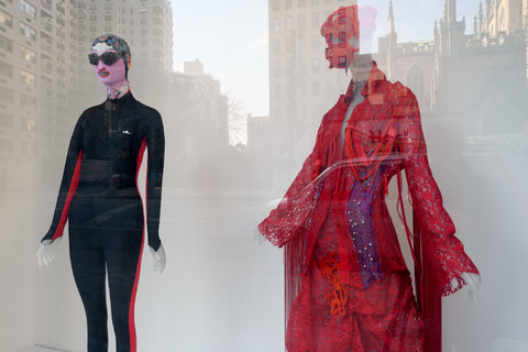 Image of two costumes from the exhibition on mannequins. On the left, a mannequin wears a black wetsuit with red stripes along side the body. It also wears goggles and a swim cap. Next to it, on the right, another mannequin wears an ornate red dress with a red bonnet-like hat.  